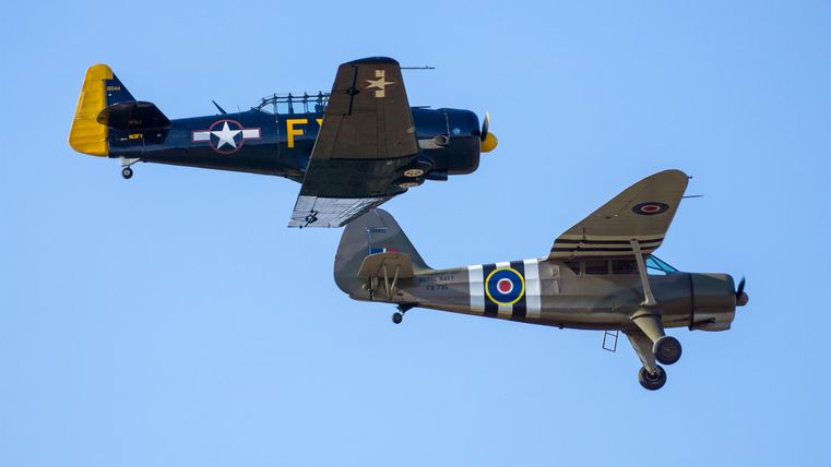 Zwei alte Flugzeuge fliegen in der Luft. Das obere Flugzeug hat eine gelbe und schwarze Lackierung, während das untere Flugzeug eine grüne und weiße Farbe hat.