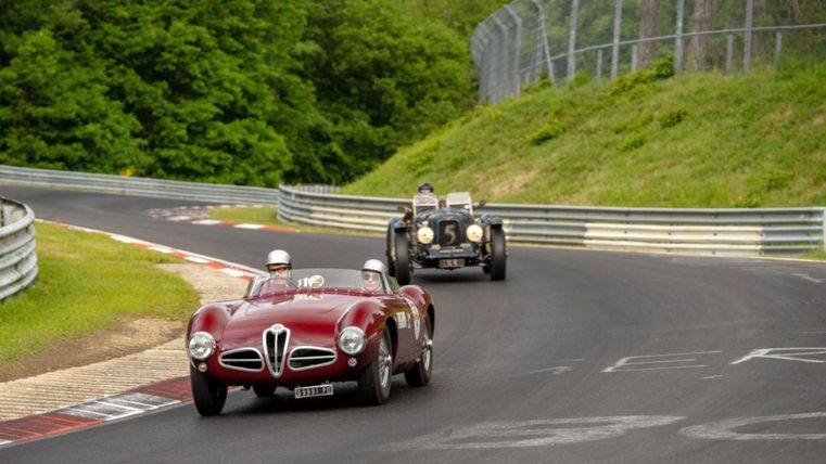 Zwei klassische Autos fahren schnell auf einer kurvenreichen Rennstrecke. Die Umgebung ist grün und voller Bäume.