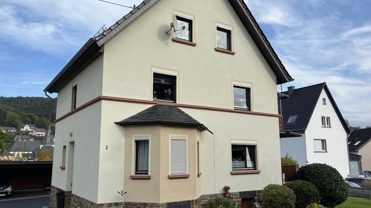 Une maison d'habitation de trois étages aux couleurs vives avec un jardin bien entretenu. En arrière-plan, d'autres bâtiments et des collines douces sont visibles.