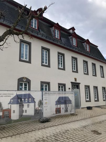 A historical building with a red roof and white walls. In the foreground, there is a construction fence featuring a visualization of the future appearance.