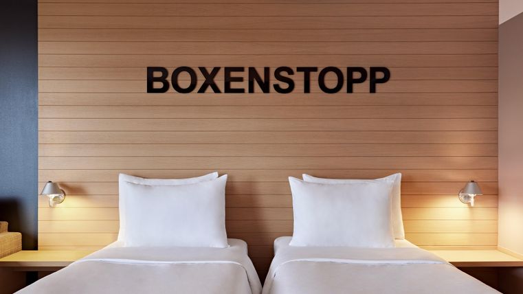 A modern hotel room with two single beds. On the wall is the inscription "BOXENSTOPP".