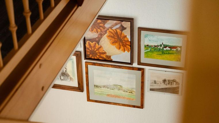 Eine Treppe, die auf mehrere Bilder an der Wand zeigt. Die Bilder zeigen unter anderem die Dorfansicht und Stillleben in Rahmen.