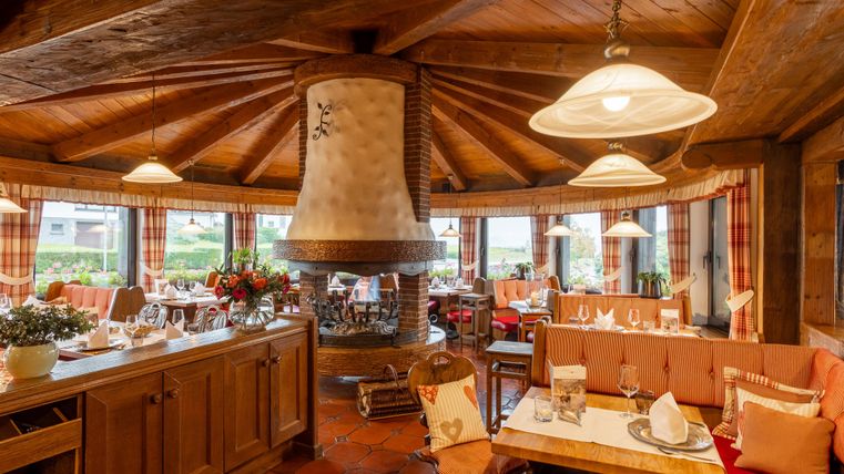 Ein gemütliches Restaurant mit Holzdecke und einem zentralen Kamin. Große Fenster bieten einen Blick ins Freie und sorgen für eine freundliche Atmosphäre.