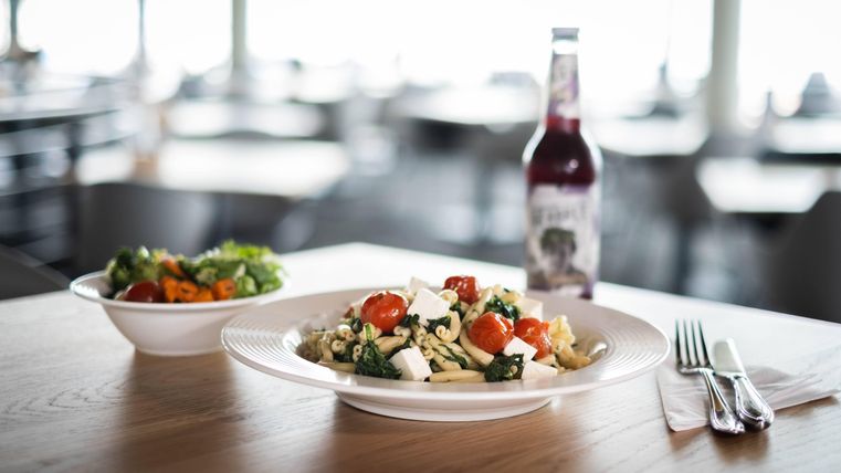 Ein Teller Pasta mit frischen Tomaten und Feta-Käse liegt auf einem Tisch. Daneben befindet sich eine Schüssel mit gemischtem Salat und eine Flasche Getränk.