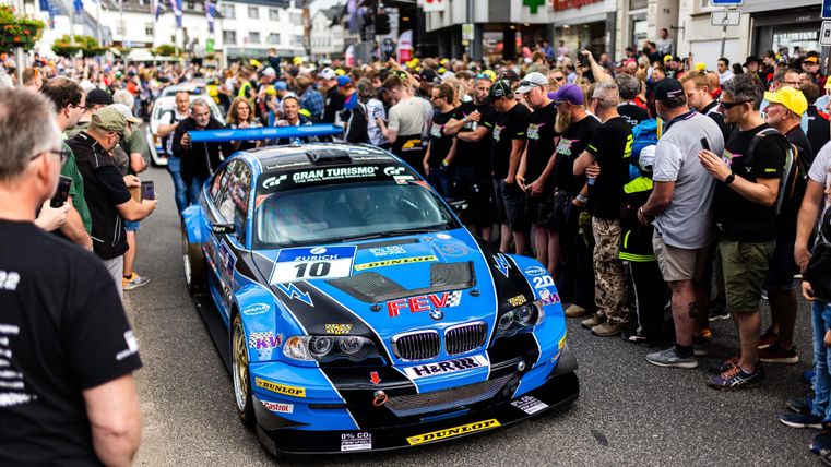 Ein Rennwagen steht vor einer großen Menschenmenge auf der Straße. Die Zuschauer zeigen Begeisterung und warten auf das Rennen.