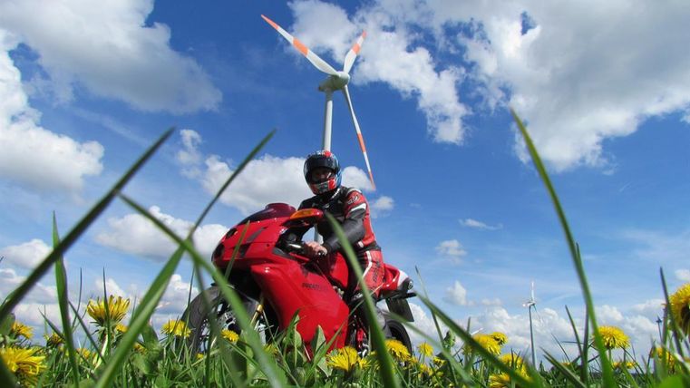 A motorcyclist is standing on a red machine in a meadow with green grass and yellow flowers. In the background, wind turbines can be seen under a blue sky with clouds.