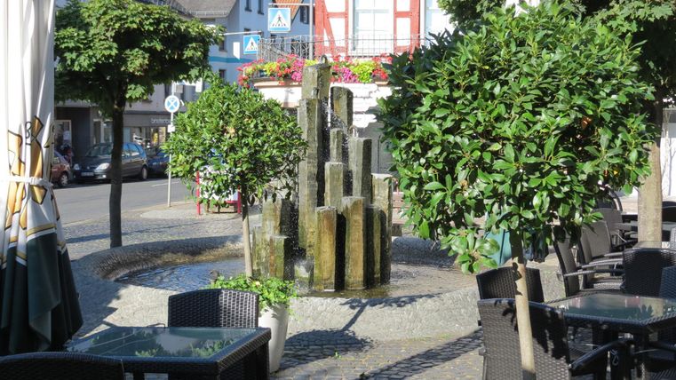 A beautiful place with a modern fountain and well-maintained trees. Around it are tables and chairs for relaxing outdoors.