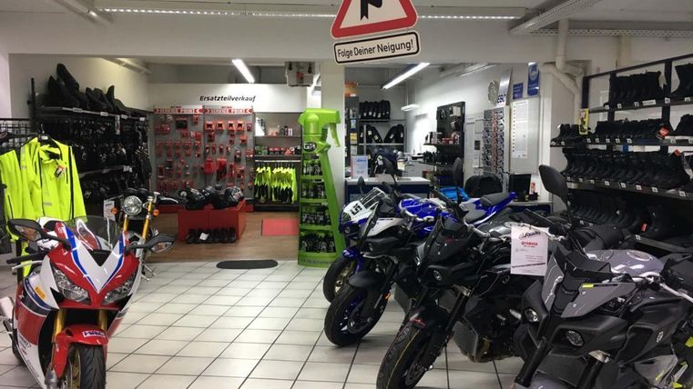 Ein Motorradgeschäft mit mehreren Motorrädern in verschiedenen Farben. Im Hintergrund sind Motorradjacken und Zubehör zu sehen.