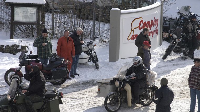 A group of people stands in the snow while motorcycles pass by. In the background, a large sign of the campsite can be seen.