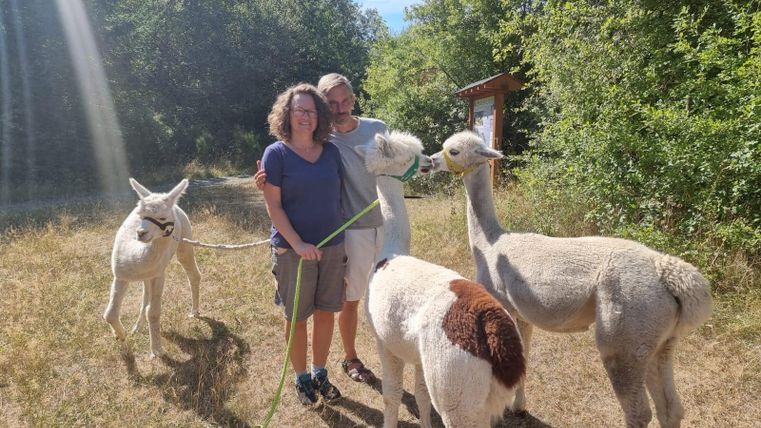 Ein Paar steht in der Natur und lächelt, während sie zwei Alpakas an Leinen halten. Im Hintergrund sind Bäume und ein blauer Himmel zu sehen.