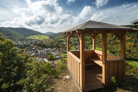 Ein Holzpavillon bietet einen Ausblick auf ein malerisches Tal und ein kleines Dorf. Im Hintergrund sind sanfte Hügel und ein klarer Himmel zu sehen.