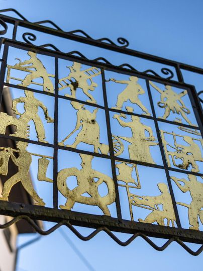 Ein kunstvoller Metallrahmen mit goldenen Figuren, die verschiedene Aktivitäten darstellen. Der Hintergrund ist ein klarer blauer Himmel.