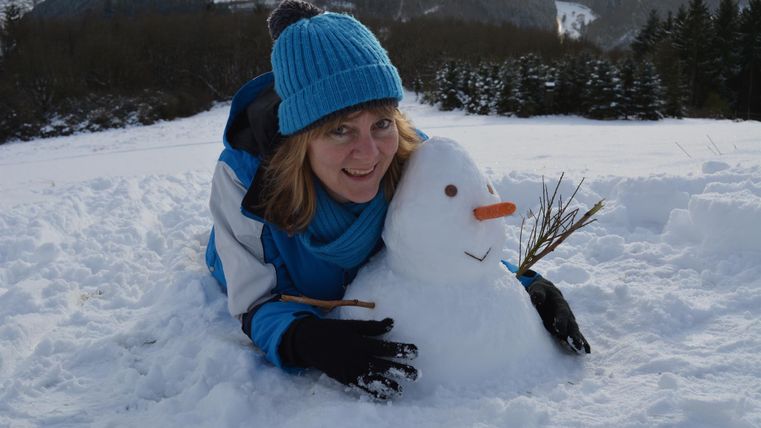 Eine Frau liegt im Schnee und posiert neben einem Schneemann. Sie trägt einen blauen Hut und einen blauen Winteranzug.