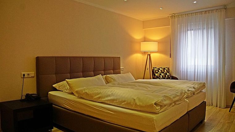 Une chambre moderne avec un grand lit double et un coin salon confortable. La pièce a une lumière chaude et de grandes fenêtres avec des rideaux.