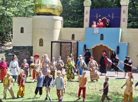 Een levendige scène met veel kinderen en volwassenen in kleurrijke kostuums. Op de achtergrond is een groot, fantasierijk gebouw met een gouden koepel te zien.
