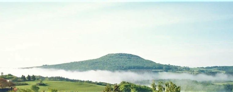 Een zachte, groene landschap met een berg op de achtergrond. Dunne mist zweeft over de vlakte onder een heldere lucht.