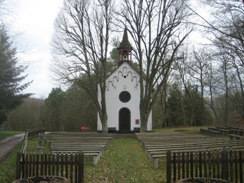 Een kleine, wit geverfde kapel met een toren staat in het midden van bomen. Voor de kapel zijn zitbanken geplaatst, die zich in een rustige omgeving bevinden.