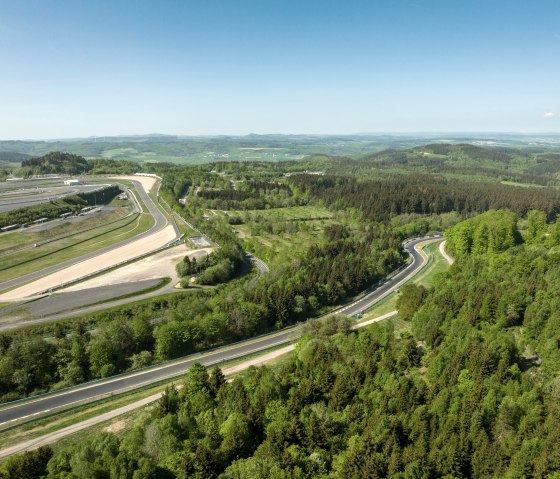 Luftaufnahme des N&uuml;rburgrings, umgeben von gr&uuml;nen W&auml;ldern und H&uuml;geln. Die Rennstrecke schl&auml;ngelt sich durch die Landschaft unter blauem Himmel., &copy; TI Hocheifel-N&uuml;rburgring, Dominik Ketz