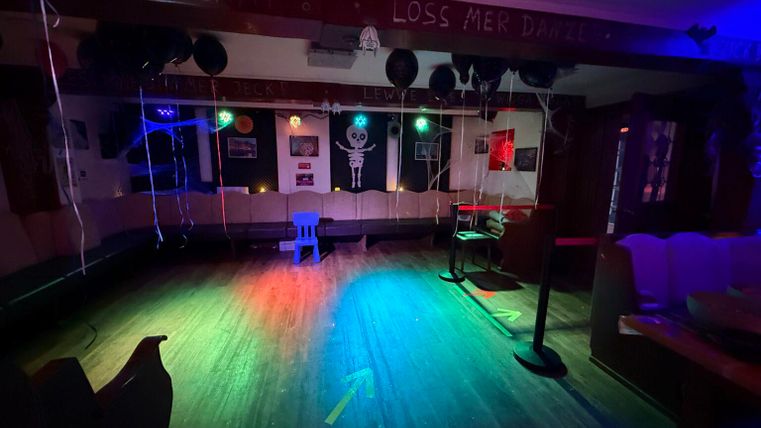 Een lege evenementenruimte met kleurrijke lichten en een houten vloer. Aan de muren hangen decoratieve elementen, waaronder ballonnen en kunstwerken.