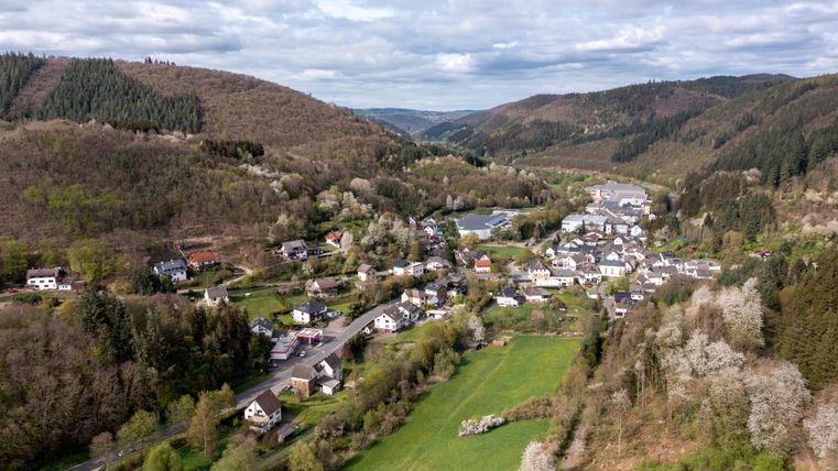 Eine malerische Landschaft mit einem kleinen Dorf, umgeben von sanften Hügeln und Bäumen. Ein Fluss schlängelt sich durch die grüne Wiese im Vordergrund.