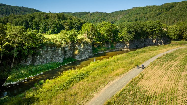 A picturesque river flows through a green landscape. Along a path, two people are riding their bikes.