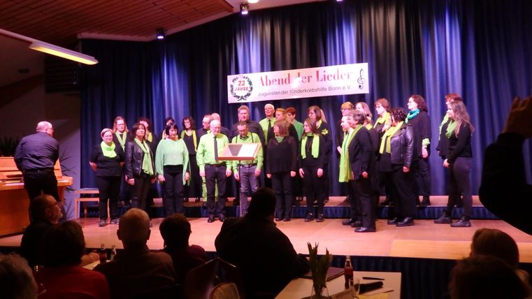 Ein Chor in bunten Outfits singt auf einer Bühne. Im Publikum sitzen viele Zuhörer, die die Darbietung genießen.