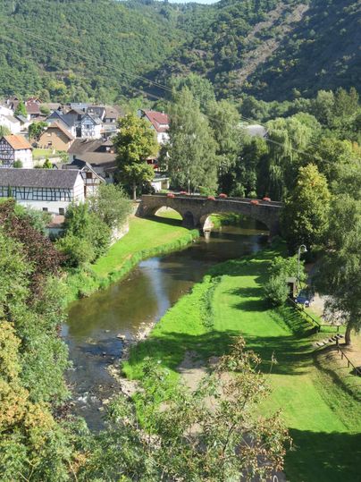 Eine malerische Landschaft mit einem Fluss, umgeben von Bäumen und einer kleinen Brücke. Im Hintergrund sind das Dorf und sanfte Hügel zu sehen.