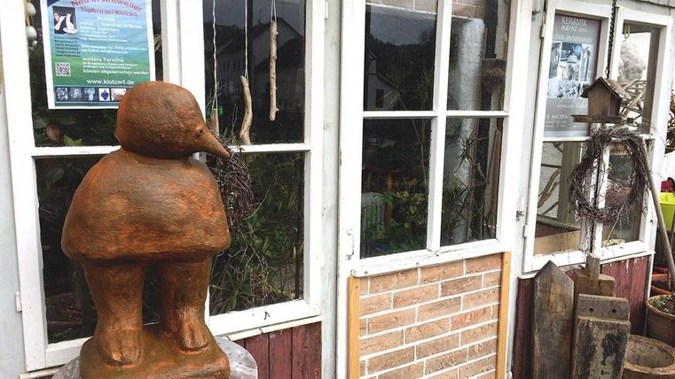 Een creatieve terracotta-sculptuur staat voor een raam. Op de achtergrond zijn planten en decoratieve elementen zichtbaar.