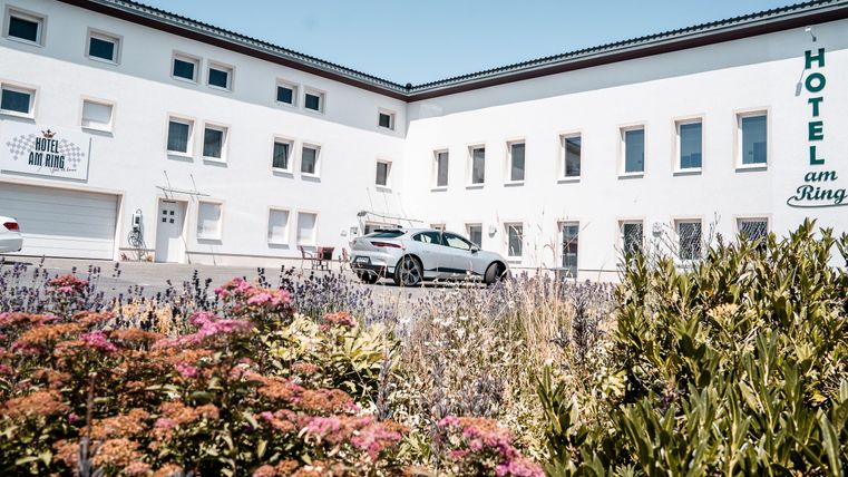 Ein modernes Hotelgebäude mit einem schönen Garten und bunten Blumen. Im Vordergrund ist ein Parkplatz mit einem Auto sichtbar.