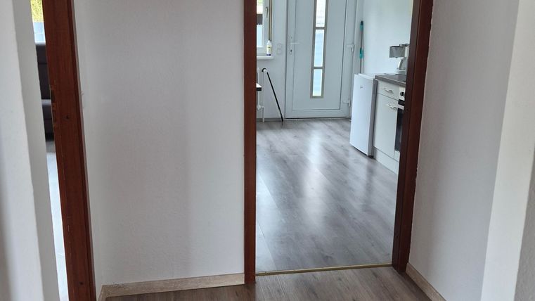 A bright hallway with wooden flooring and two doors. In the background, a modern kitchen is visible.