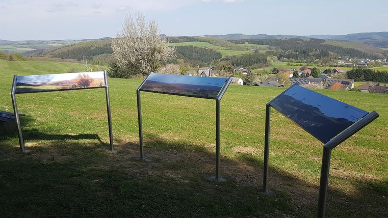 Drei Informationsschilder stehen auf einer Wiese mit Blick auf die Landschaft. Im Hintergrund sind sanfte Hügel und ein klarer Himmel zu sehen.