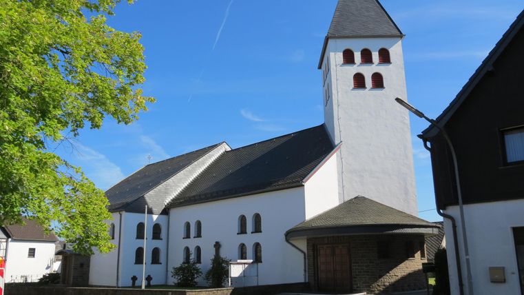 Eine Kirche mit einem charakteristischen Turm und weißen Wänden. Im Vordergrund befindet sich eine ruhige Straße und grüne Bäume.