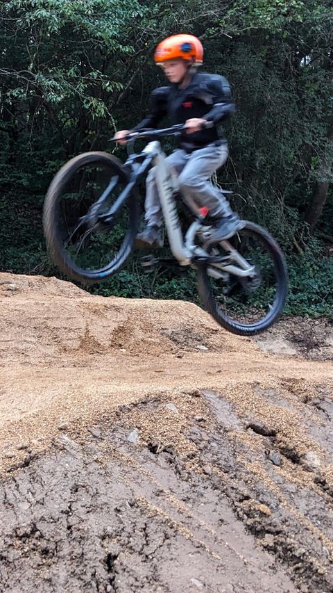 Ein junger Radfahrer springt mit seinem Mountainbike über einen Hügel aus Sand. Er trägt einen orangefarbenen Helm und hat eine sportive Ausrüstung.