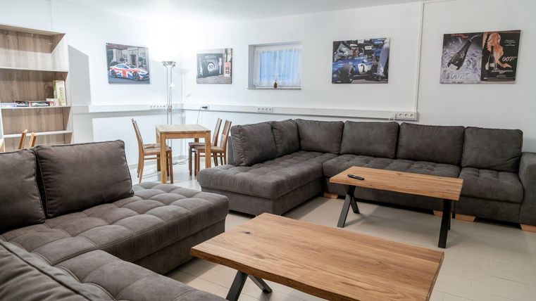 Een moderne woonkamer met een grote, grijze bank en houten tafels. Aan de muren hangen verschillende foto's en er is een eettafel met stoelen.