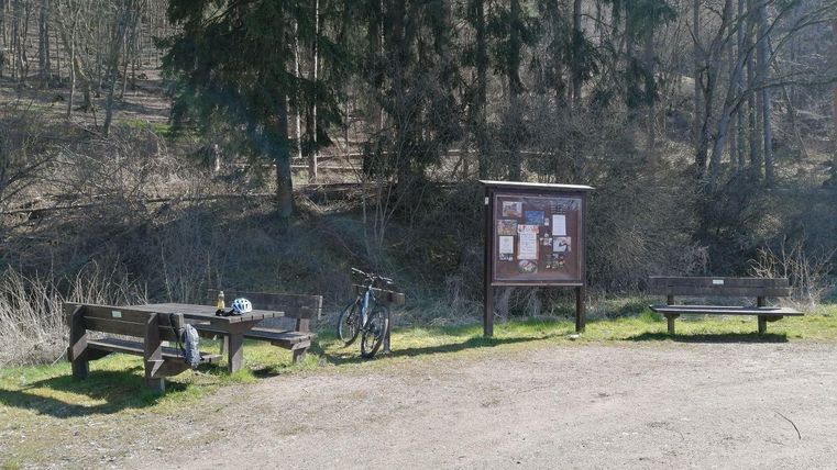 Eine gemütliche Raststätte im Wald mit Holzbänken und einem Informationsschild. Ein Fahrrad steht neben den Bänken.