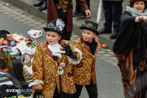 Zwei Kinder in auffälligen Kostümen mit Zylinderhüten stehen auf einer Straße. Sie posieren fröhlich und genießen eine festliche Umgebung.