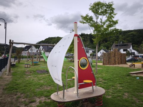 Ein Spielplatz mit einer Segelboot-Spielanlage und einem runden Fenster. Im Hintergrund sind Bäume und Wohnhäuser zu sehen.