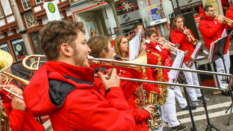 Eine Blaskapelle spielt auf einer Straße. Die Musiker tragen rote Jacken und sind konzentriert bei der Aufführung.