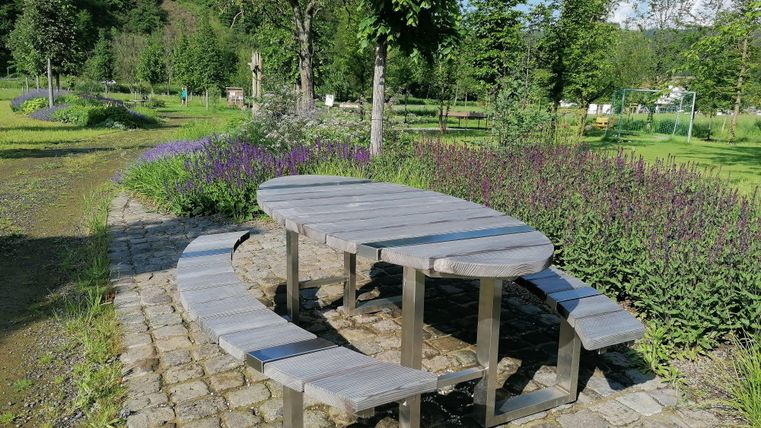 Ein runder Tisch mit Bänken steht in einem Garten. Umgeben von Bäumen und Blumen bietet der Platz eine einladende Atmosphäre.