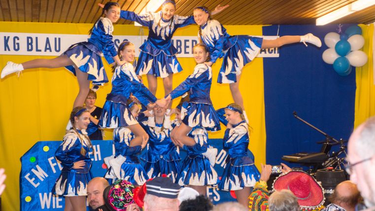 Een groep dansers in blauwe kostuums presenteert een acrobatische vertoning. De omgeving is feestelijk met kleurrijke versieringen.