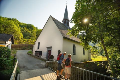 Eine kleine Kirche mit einem spitzen Turm in einer idyllischen Landschaft. Drei Wanderer stehen auf einer Brücke und genießen die Aussicht.