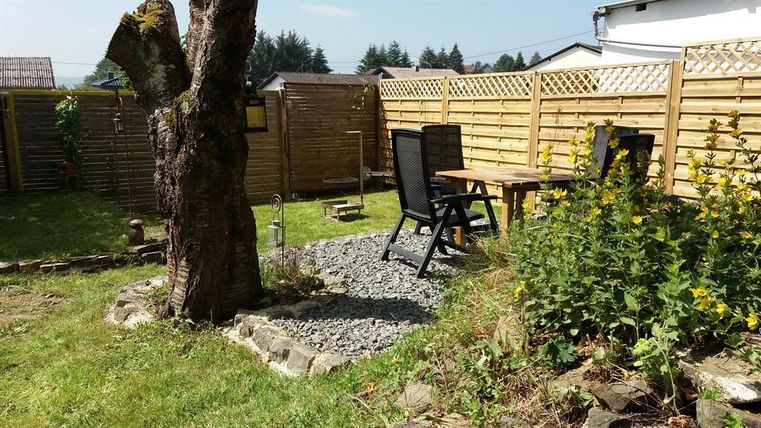 Ein Garten mit einem großen Baum, einem Kiesbereich und einer gemütlichen Sitzecke. Umgeben von einem Holzzaun und bunten Blumen.