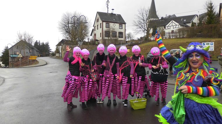 Een groep mensen in kleurrijke kostuums en roze hoeden staat vrolijk op straat. Op de achtergrond zijn enkele huizen en bomen te zien.