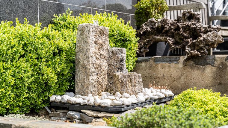 Een decoratieve steenskulptuur staat in een verzorgd tuin. Groene struiken en kiezelstenen omlijsten de scène.