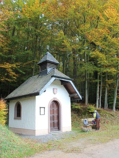 Kleine Kapelle im Wald mit einer Person auf einer Bank daneben.