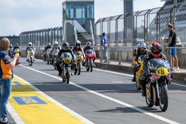 Eine Gruppe von Motorrädern fährt auf der Rennstrecke. Zuschauer beobachten das Geschehen von der Seite aus.