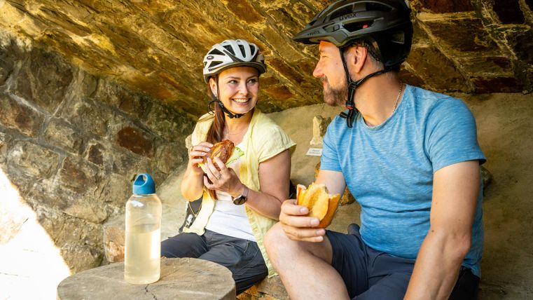 Ein Paar sitzt in einer gemütlichen Ecke und genießt ihr Essen. Sie tragen Fahrradhelme und haben eine Wasserflasche neben sich.