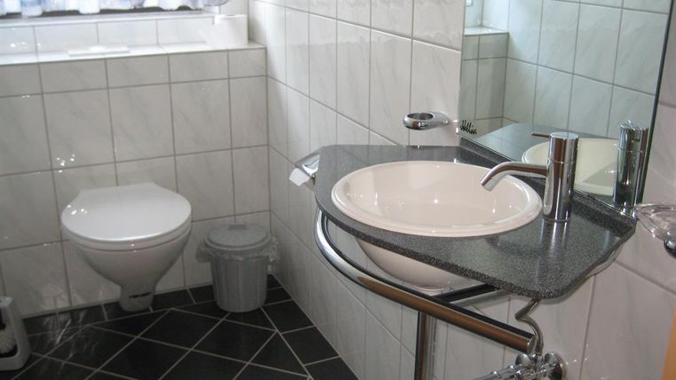 Une salle de bain moderne avec des carreaux blancs et un lavabo rond. À gauche se trouve une toilette et une poubelle.