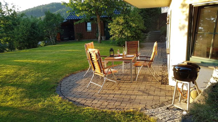 Ein gemütlicher Garten mit einem runden Tische und Stühlen. Im Hintergrund sind Bäume und ein Holzhaus sichtbar.