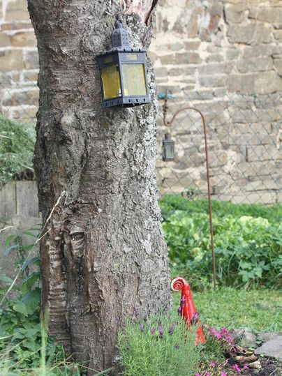 Ein alter Baum mit einer Laterne hängt daran. Im Vordergrund blühen bunte Blumen und eine rote Figur steht in der Nähe.
